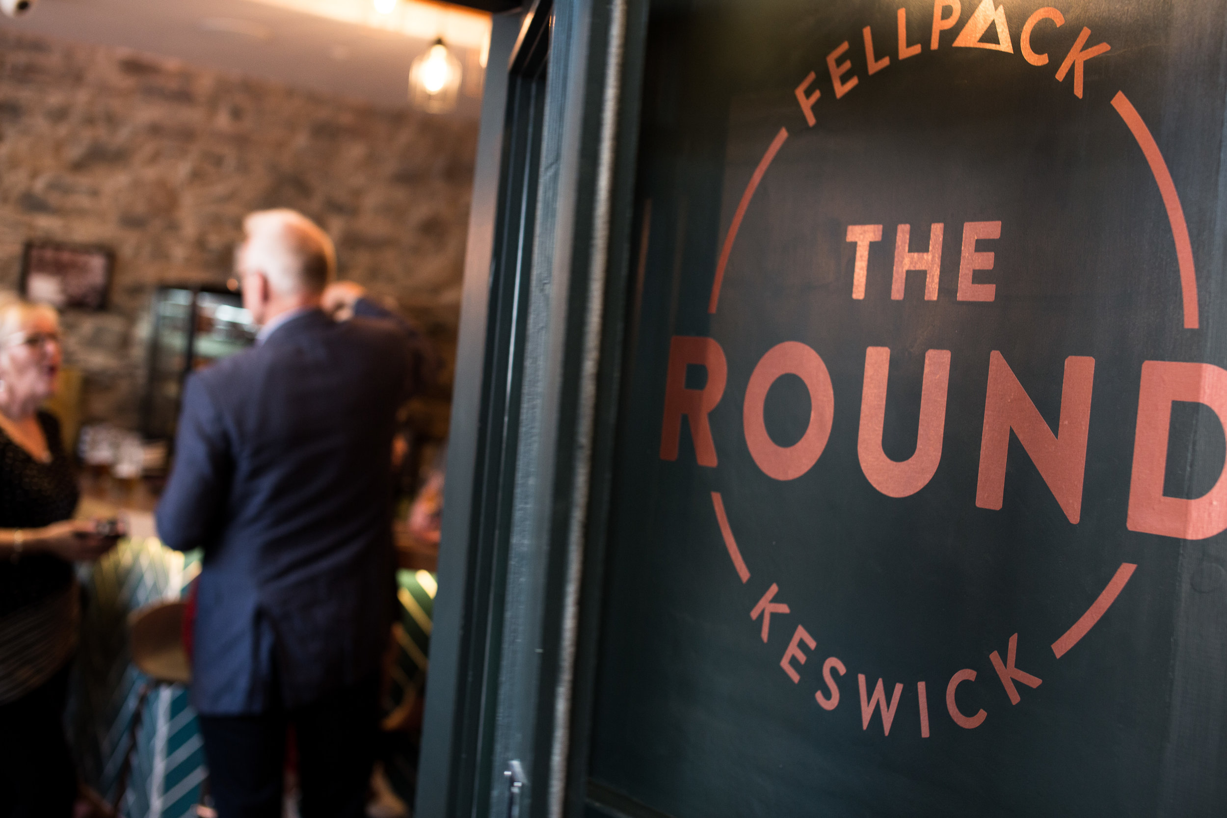 The image shows the interior sign on the entrance into the Round, Keswick's most popular burger bar. Out of focus, next to the sign, is a man in a suit and a woman speaking to him.